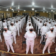 Soldados de pé, em filas, ao lado de suas cadeiras, com farda branca durante o evento de nomeação.