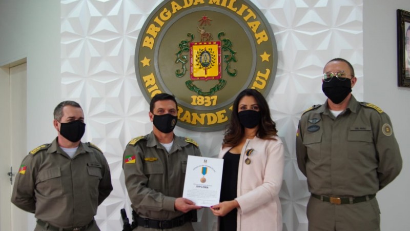 Diante de uma parede com brasão da BM, coronel Stumf, coronel Santarosa e a jornalista Zete Padilha, esses dois últimos segurando um papel de certificado, e ainda o coronel Feoli.