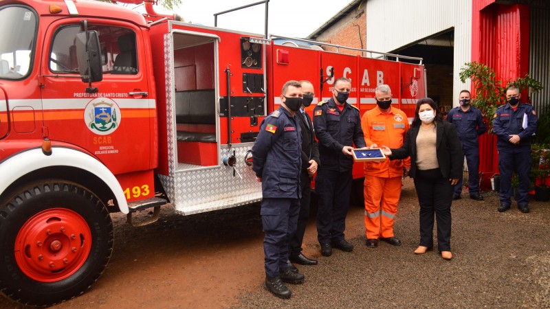 Em frente ao caminhão de bombeiros, vermelho e estilizado com o Layout di SCAB, os bombeiros militares e autoridades da cidade entregam documento de doação de caminhão de bombeiros para a os bombeiros voluntários