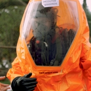 Um dos bombeiros está utilizando um traje para o manuseio de produtos perigosos.