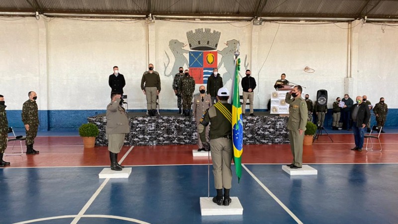 Cerimônia de posse do Comandante dentro de um galpão esportivo. Os integrantes estão nas posições designadas. Temos a presença de um palco onde se apresentam 7 pessoas, as outras pessoas estão na parte de baixo.