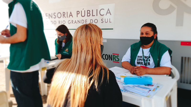 Mulher sentada recebendo atendimento por servidora da defensoria pública, também sentada, do lado oposto da mesa de plástico branco, elas estão na sobra de um toldo que está preso a um ônibus da defensoria pública.