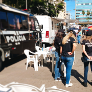 Três integrantes da Polícia Civil, com camiseta e boné preto da instituição, de pé em frente a mesas e cadeiras de plásticos que estão dispostas ao lado de um ônibus também da Polícia Civil.