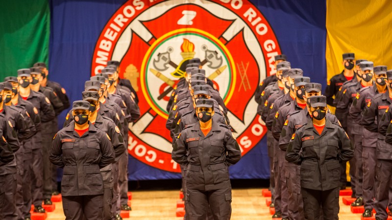 Cinco filas de bombeiros lado a lado em posição descansar. Ao fundo, pano azul com o brasão do CBMRS.