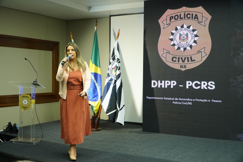 Delegada Vanessa Pitrez, em pé, de vestido marrom e casaco bege, fala ao microfone. Ao lado esquerdo, um púlpito com símbolo da PC. Ao fundo, bandeiras do Brasil e da PC. À direita, telão com brasão da PC e o nome DHPP - PC.