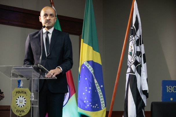 Delegado Fábio Motta Lopes de terno preto, gravata preta e camisa branca, em pé, diante de um púlpito, fala ao microfone. Ao lado, bandeiras do Brasil e da Polícia Civil.