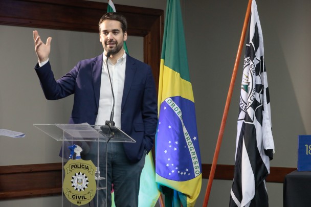 Governador Eduardo Leite, de casaco azul e camisa branca, em pé, diante de um púlpito, fala ao microfone. Ao lado, bandeiras do Brasil e da Polícia Civil.