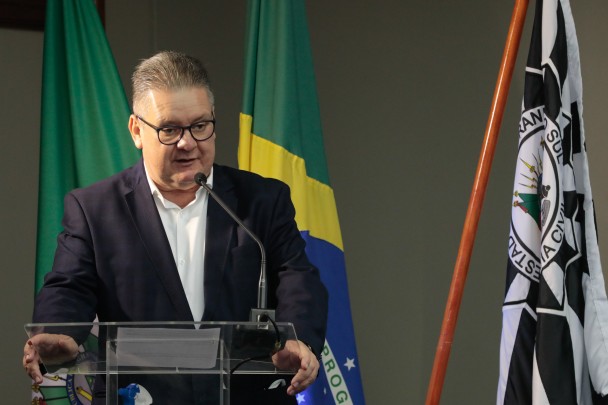 Delegado Ranolfo, de óculos, terno preto e camisa branca, em pé, fala ao microfone. Ao fundo, bandeiras do Brasil e da PC.