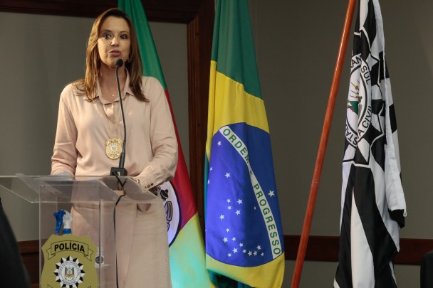 Delegada Nadine Anflor, em pé, de camisa e saia beges, com distintivo de delegada pendurada no pescoço, fala ao microfone diante de um púlpito. Ao lado, bandeiras do RS, do Brasil e da Polícia Civil.