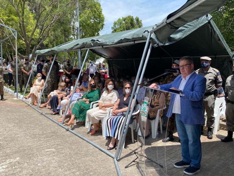 Delegado Ranolfo, de sapato preto, calça jeans, camisa branca e paletó azul, em pé, discursa ao microfone diante de um púlpito. Ao fundo, à esquerda, pessoas sentadas em cadeiras sob tendas metálicas com lona verde do Exército.