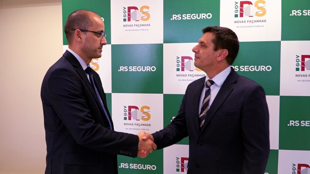 Delegado Fábio Motta Lopes, em pé, de terno e gravata, aperta a mão do secretário da Segurança Pública, coronel Vanius Cesar Santarosa, também em pé, de terno de gravata. Ao fundo, banner quadriculado com as logos do governo e do RS Seguro.
