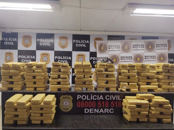 Sobre uma bancada com a inscrição "Polícia Civil 08000518518 Denarc", 197 tijolos marrons de maconha dispostos em pilhas lado a lado. Em cima de uma das pilhas, ao centro da imagem, um revólver calibre 38. Ao fundo, banners quadriculados da Polícia Civil e da Brigada Militar.