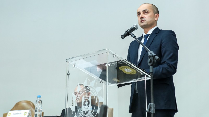 Delegado Fábio Motta Lopes, de terno e gravata, em pé, fala em um microfone em um pedestal diante de um púlpito de acrílico transparente com símbolo da Polícia Civil.
