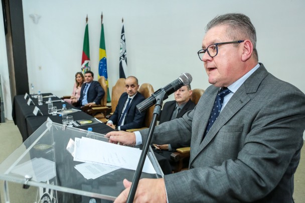 Governador Ranolfo, de terno e gravata, em pé, fala ao microfone diante de um púlpito de acrílico. Ao fundo, sentados diante de uma mesa com toalhda preta, o novo subchefe da PC, Vladimir Urach, o novo chefe, Fábio, o secretário da SSP, Santarosa, e a delegada Nadine Anflor. Mais ao fundo, bandeiras do RS, do BR e da PC.