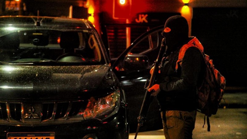 Homem segurando uma arma longa, usando balaclava e carregando um mochila às costas aparece parado, em pé, ao lado de uma camionete preta.