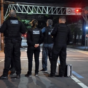 Agentes da Polícia Civil aparecem parados de costas em uma rua de Caxias do Sul. Ao lado deles, no chão, uma maleta.