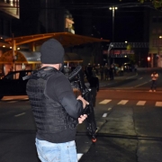 Homem armado vestindo colete e touca preta aparece parado de pé, de costas, em uma rua de Caxias do Sul. Ao fundo, no escuro, um veículo parado.