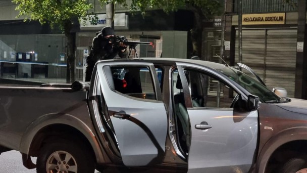 Sobre a caçamba de uma camioneta com as duas portas laterais abertas, um homem vestido de preto, inclusive com balaclava, opera uma arma longa escorada na carroceria do veículo.