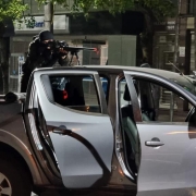 Sobre a caçamba de uma camioneta com as duas portas laterais abertas, um homem vestido de preto, inclusive com balaclava, opera uma arma longa escorada na carroceria do veículo.
