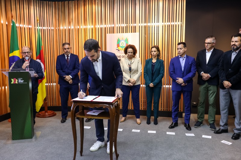 O governador Eduardo Leite, de casaco e calça azuis, camisa e tênis brancos, assinado um documento sobre um pequena mesa, em uma sala com carpete e parede de ripas de madeira. Ao fundo, outras autoridades envolvidas no projeto observação, ao lado de bandeiras do RS e do Brasil.