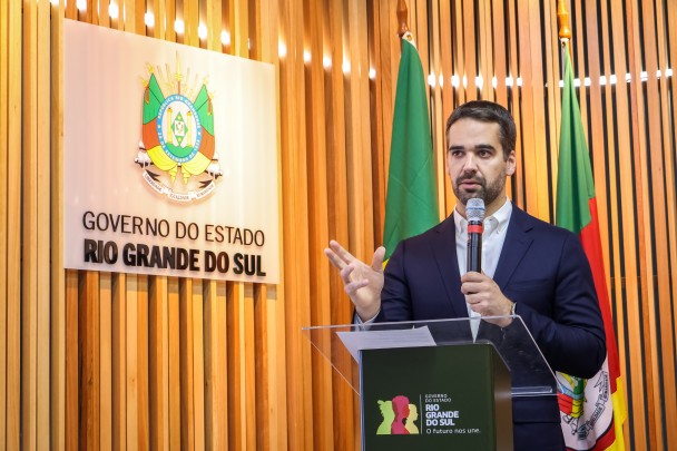 O governador Eduardo Leite fala segurando um microfone diante de um púlpito. Atrás dele, bandeiras do Brasil e do RS. Ao lado, brasão do RS em uma peça de acrílico afixada em uma parede de ripas de madeiras
