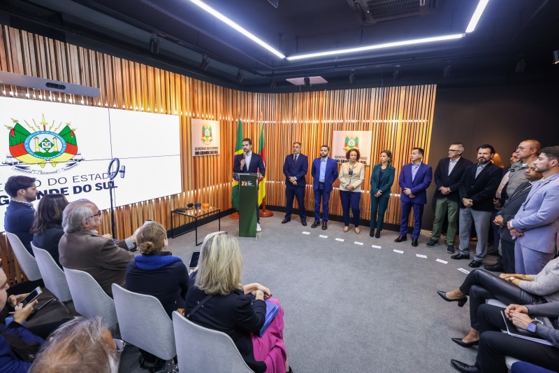 Eu uma sala com carpete cinza, o governador fala ao microfone diante de um púlpito verde. Ao lado deles, autoridades envolvidas no projeto de monitoramento do Agressor. Ao fundo, parede de ripas de madeiras com brasões do RS em peças de acrílico. À direita, telão com brasão do RS na tela. Diante das autoridades, jornalistas sentados em cadeiras acompanham o ato.