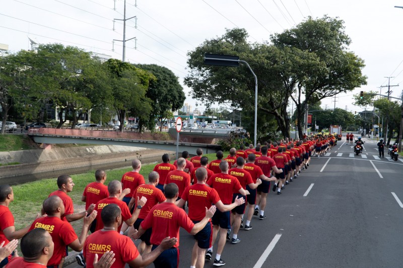 Foto mostra bombeiros praticando corrida ao longo de avenida.
