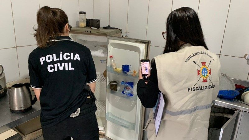 Foto mostra agentes da Polícia Civil e da Vigilância diante de geladeira