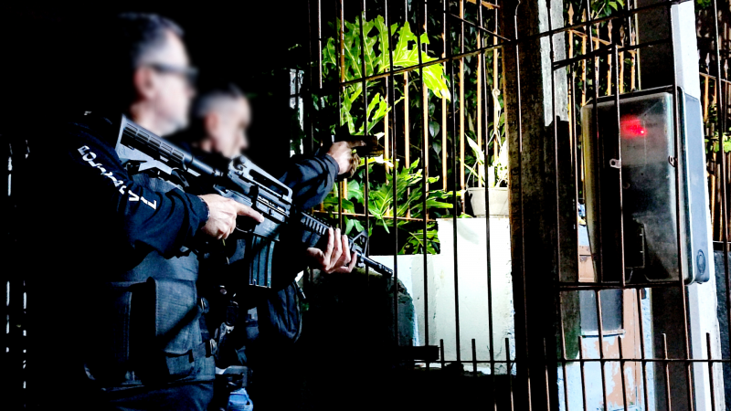dois policiais civis de perfil, empunhando armas, em frente a um portão com medidor de companhia de energia em frente
