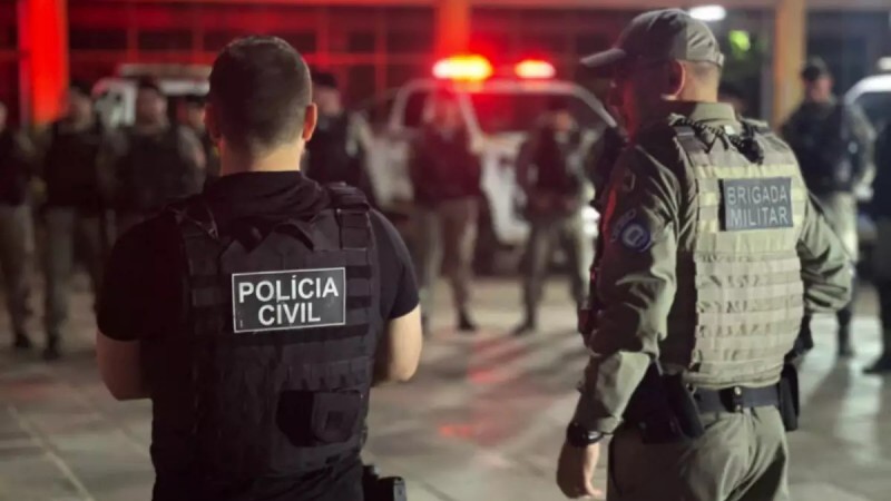 Foto mostra policial civil e brigadiano de costas lado a lado em ambiente externo. O fundo está desfocado, mas se vê policiais diante de viatura.