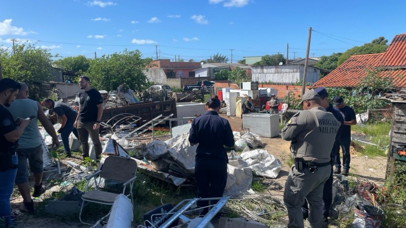 Foto em área aberta mostra agentes da PC e BM em meio a materiais de reciclagem depositados no solo.