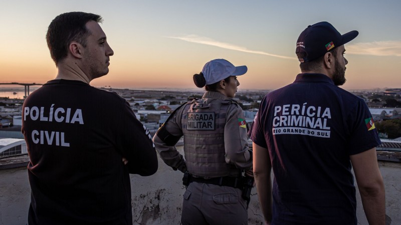 Servidores da segurança pública estão de costas na imagem: dois homens (um policial civil e um perito criminal) e uma mulher da Brigada Militar.