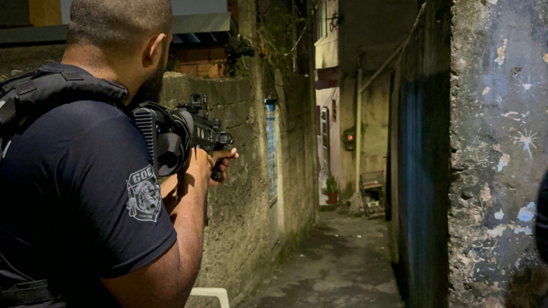 Foto mostra policial empunhando arma em ruela de casas de alvenaria.