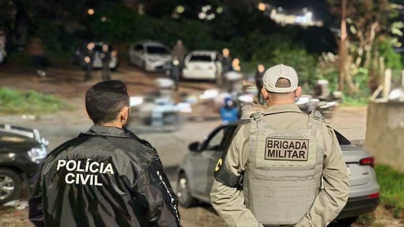 Foto externa mostra um policial civil e um policial militar lado a lado. Eles estão de costas, olhando para um local aberto com carros estacionados.