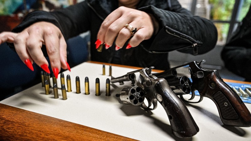 Foto mostra armas e munições sobre mesa. Uma mulher de unhas vermelhas manipula as munições.