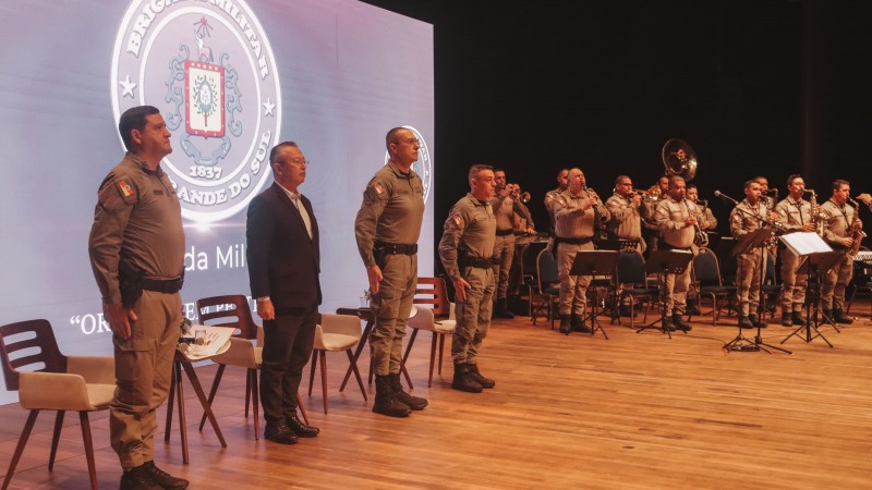 A imagem mostra oficiais e autoridades perfiladas durante execução de uma canção pela banda da Brigada Militar