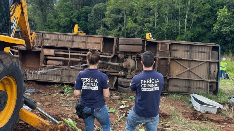 Na foto, há duas pessoas em primeiro plano, de costas, com camisetas onde se lê "Perícia Criminal Rio Grande do Sul", observando, ao fundo, um caminhão tombado. Aparecem árvores ao fundo, atrás do veículo, e há uma escavadeira amarela ao lado dos dois peritos.