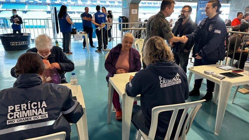 A imagem mostra uma central de atendimento à população. Há mesas brancas nas quais atendentes com uniformes do IGP (de costas) atendem populares. Atrás, há alambrados e pessoas conversando de pé e outras, sentadas.
