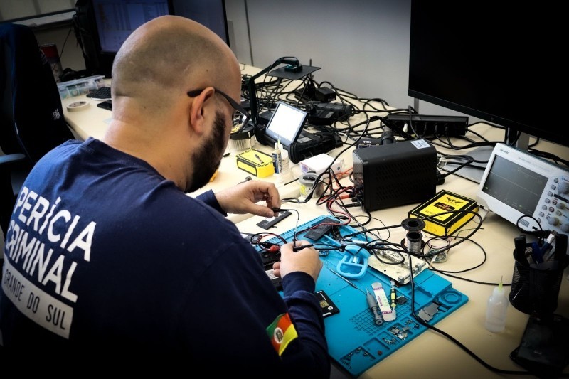 Um homem está lidando com aparelhos eletrônicos e fios dispostos sobre uma bancada. Ele está de costas e, em sua camisa, se lê: "Perícia Criminal Rio Grande do Sul".
