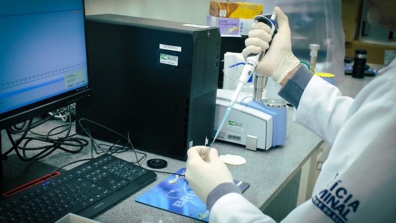 Uma pessoa não identificada (a foto não mostra o rosto), com jaleco do IGP e luvas, manuseia um instrumento na frente de um computador.