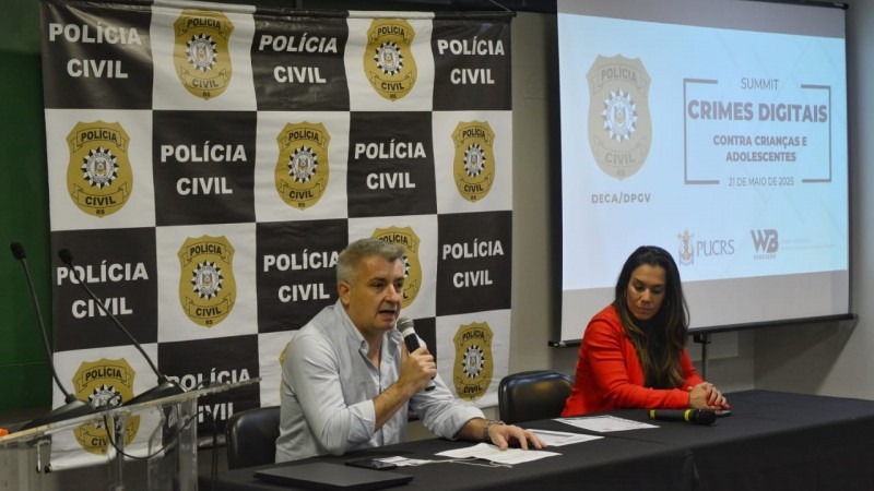 No palco do evento, especialista fala ao microfone ao lado de outra palestrante. Atrás deles, banner da Polícia Civil e uma tela de projeção onde se lê "Summit Crimes Digitais contra crianças e adolescentes" ao lado do escudo da Polícia Civil.