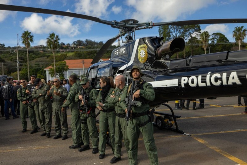 Helicóptero com identificação da Polícia Civil. À frente dele, policiais vestidos com macacões verdes, segurando armamentos.