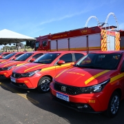 Cinco viaturas vermelhas estão à frente de dois novos caminhões do Corpo de Bombeiros Militar. Ao fundo, a tenda onde aconteceu o ato simbólico da entrega