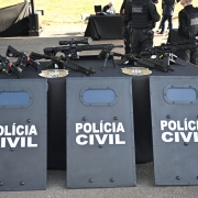 Sobre uma mesa coberta com toalha preta, estão dispostas armas e, apoiados no chão, escudos balísticos com identificação da Polícia Civil