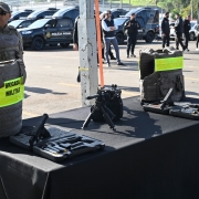 Sobre uma mesa coberta com toalha preta, dois coletes à prova de balas, duas pistolas em suas maletas de transporte e, ao centro, um fuzil.