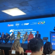 A foto, tirada da plateia, mostra o palco do local do evento. Há um telão com fundo azul sobre o qual se lê "2º Encontro Nacional do Programa de Compras Eficientes para o SUSP". À frente, há oito pessoas sentadas, voltadas para o público, e uma mulher fala no púlpito.