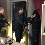Policiais civis vasculham cômodo em uma casa. Panelas e outros utensílios de cozinha estão sobre uma mesa. Agentes fardados levantam uma cortina para acessar o cômodo anexo. 