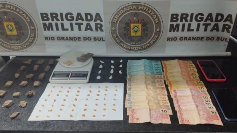 A imagem mostra drogas, balança de precisão, dinheiro e celulares apreendidos sobre uma mesa. Atrás há um banner da Brigada Militar