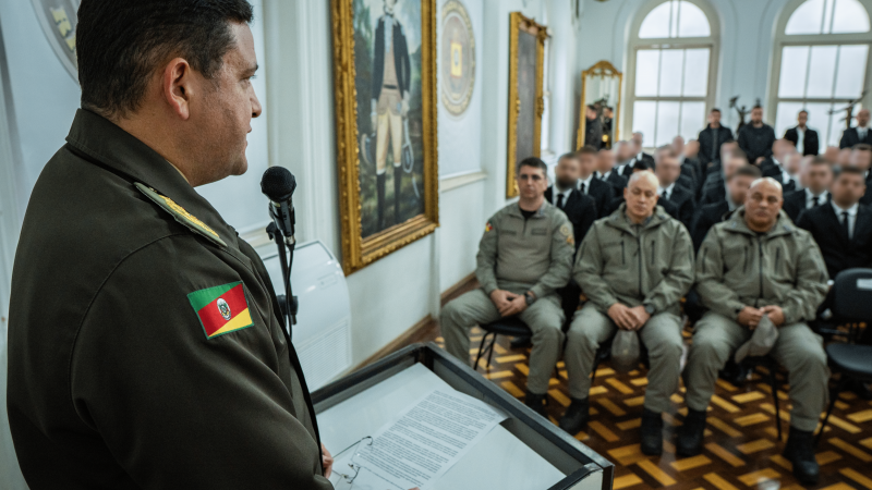 A imagem mostra um oficial da Brigada Militar discursando em um púlpito para uma plateia composta por policiais militares e formandos da 1ª Edição do Curso de Especialização em Inteligência Policial