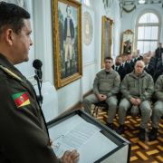 A imagem mostra um oficial da Brigada Militar discursando em um púlpito para uma plateia composta por policiais militares e formandos da 1ª Edição do Curso de Especialização em Inteligência Policial